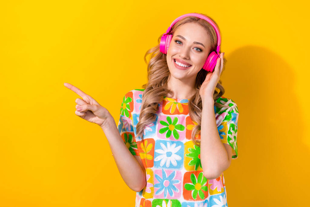 Portrait of satisfied woman wear flower print t-shirt indicating empty space touching headphones isolated on yellow color background. - Foto, Imagen