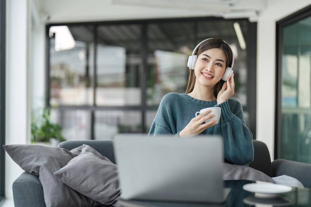 Sonriendo mujer asiática en los auriculares usando ordenador portátil mientras tiene su café de la mañana sentado en un sofá en casa. - Foto, Imagen