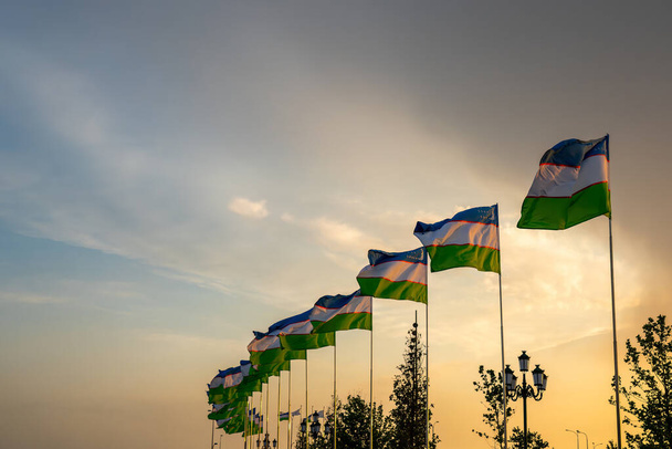 Flags of Uzbekistan waving on a sunset or sunrise dramatic cloudy sky background. - Фото, изображение