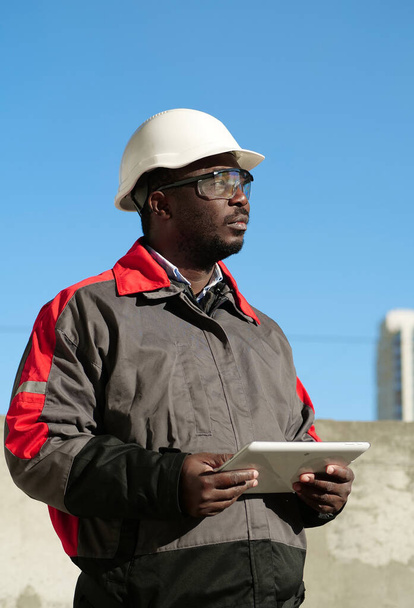 Αφροαμερικανός εργαζόμενος με σκληρό καπέλο και προστατευτικά γυαλιά με tablet υπολογιστή στέκεται στο εργοτάξιο - Φωτογραφία, εικόνα