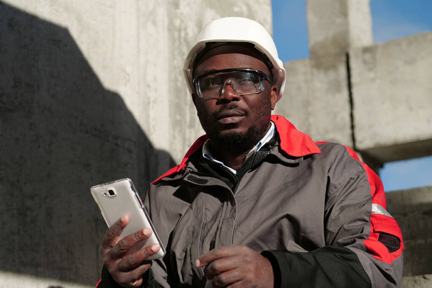 Trabajador afroamericano en sombrero duro y gafas protectoras con teléfono inteligente se sienta en el sitio de construcción. Workman sostiene en las manos el teléfono inteligente blanco y piensa - Foto, Imagen