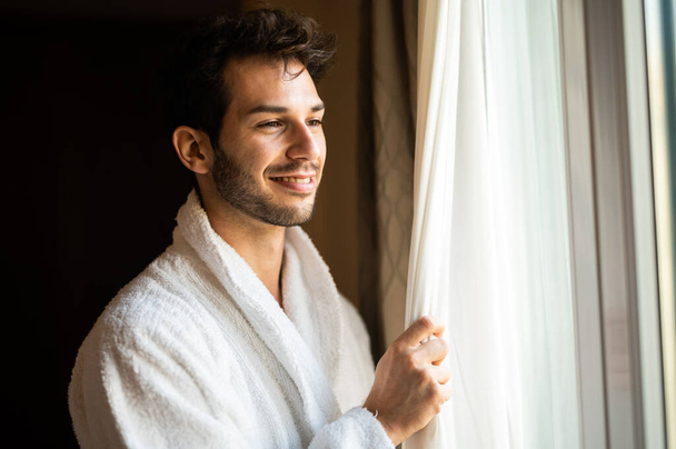 Jovem vestindo um roupão de banho e olhando para fora de uma janela em seu quarto de hotel - Foto, Imagem
