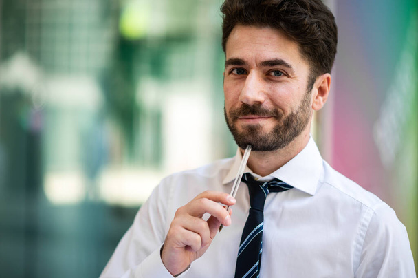 Young businessman outdoor smiling while holding a pen under his chin - Φωτογραφία, εικόνα