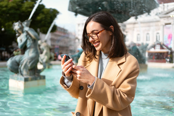 Γυναίκα φορώντας γυαλιά, κάθεται στο σιντριβάνι, κρατώντας smartphone και χαμογελώντας γλυκά. Καλές δονήσεις και αισθήματα. Ηλεκτρονική επικοινωνία με χρήση κινητής ψηφιακής συσκευής - Φωτογραφία, εικόνα