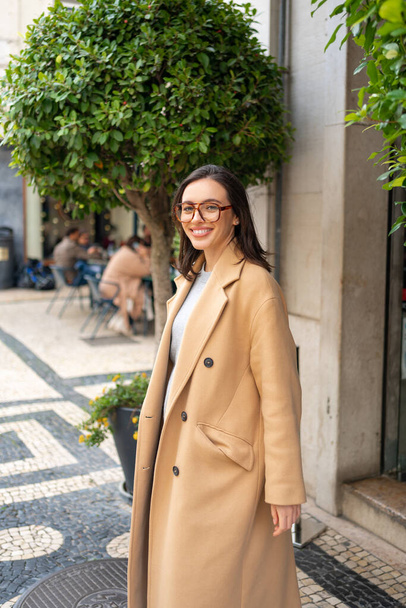Přátelsky se usmívající atraktivní žena opouští budovu na pozadí zeleného plotu v ulici evropského města. Šťastná sebevědomá žena v brýlích oblečené stylové trenč kabát s úsměvem při pohledu do kamery - Fotografie, Obrázek