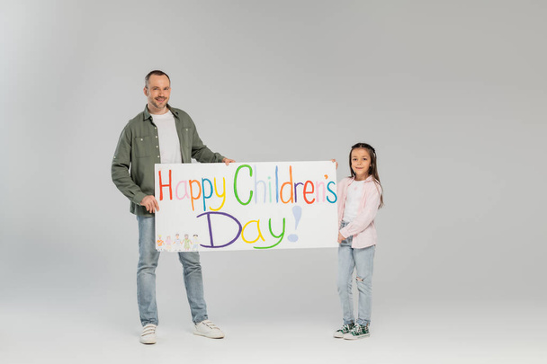 Full length of smiling father and preteen daughter in casual clothes looking at camera while holding placard with happy children's day lettering while celebrating together on grey background - 写真・画像