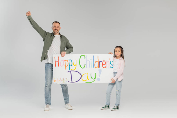 Smiling preteen girl in casual clothes holding placard with happy children's day lettering near father showing yes gesture and looking at camera on grey background - Foto, afbeelding