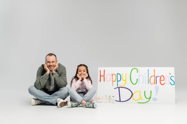 Smiling father and preteen daughter in casual clothes looking at camera while sitting near placard with happy children's day lettering during celebration in June on grey background - Foto, Bild