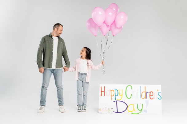 Full length of smiling preteen girl in casual clothes holding festive balloons and hand of father near placard with happy children's day lettering on grey background - Fotó, kép