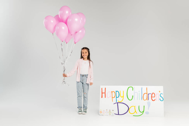 ピンクの風船を持っているカジュアルな服の中で笑顔のpreteenの子供の完全な長さとグレーの背景でお祝いの間に幸せな子供の日のレタリングとプラカードの近くのカメラを見て - 写真・画像