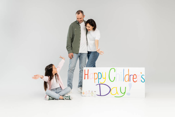 Opgewonden ouders kijken naar een kleine dochter in casual kleding zitten in de buurt van bordje met gelukkige kinderen dag belettering tijdens de viering in juni op grijze achtergrond - Foto, afbeelding