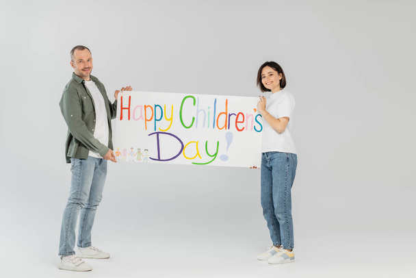Full length of smiling adult couple in casual clothes looking at camera while holding placard with happy children's day lettering together while standing on grey background - Photo, Image