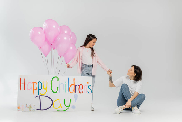 Mère tatouée souriante tenant la main de la fille préadolescente près d'une pancarte avec des lettres d'enfants heureux et des ballons roses pendant la célébration en juin sur fond gris - Photo, image