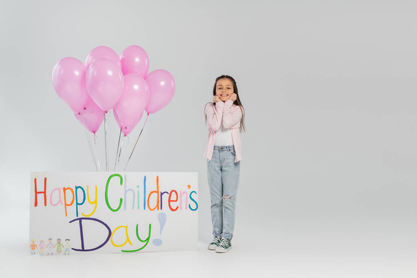 Πλήρες μήκος ανέμελη preteen κορίτσι σε casual ρούχα κοιτάζοντας κάμερα, ενώ στέκεται κοντά σε ροζ μπαλόνια και πλακάτ με γράμματα ευτυχισμένη ημέρα των παιδιών σε γκρι φόντο - Φωτογραφία, εικόνα