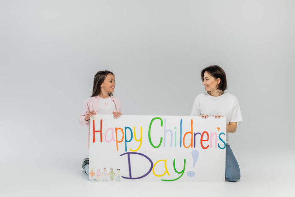 Smiling preteen girl looking at brunette and positive mother near placard with happy children's day lettering during celebration in June on grey background with copy space  - Fotó, kép