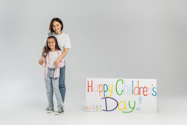 Adult woman in casual clothes hugging cheerful preteen daughter and looking at camera near placard with happy children's day lettering during celebration on grey background - Foto, immagini