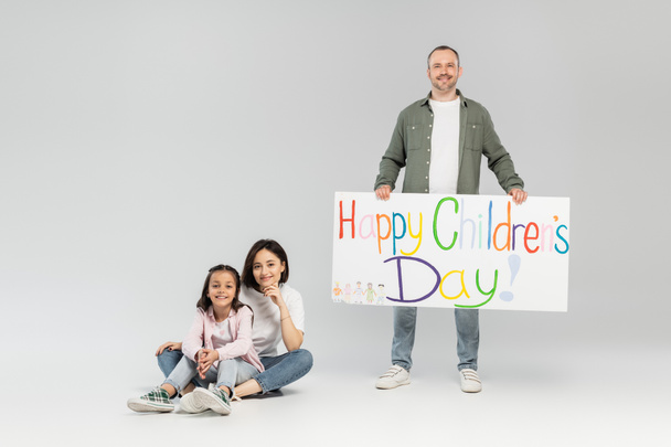 Full length of smiling father holding placard with happy children's day lettering near wife and preteen daughter looking at camera while sitting and celebrating in June on grey background - Foto, immagini