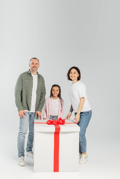 Full length of smiling parents and preteen daughter looking at camera near big gift box during child protection day celebration on grey background - Photo, Image