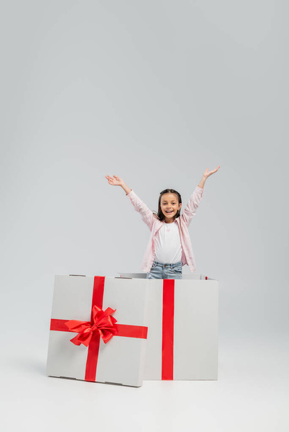 Excited and cheerful preteen girl in casual clothes looking at camera while standing in big present box during international children day on grey background - Foto, Imagen