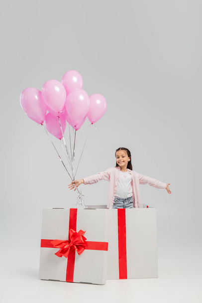 Carefree preteen kid in casual clothes looking at camera while holding pink festive balloons while standing in big present during international children day celebration on grey background - Fotó, kép