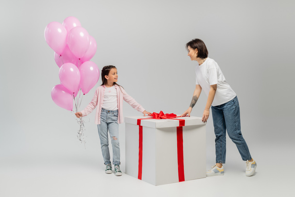 Full length of smiling preteen girl holding pink balloons and looking at tattooed mom near big present box during happy children day celebration on grey background - Photo, Image