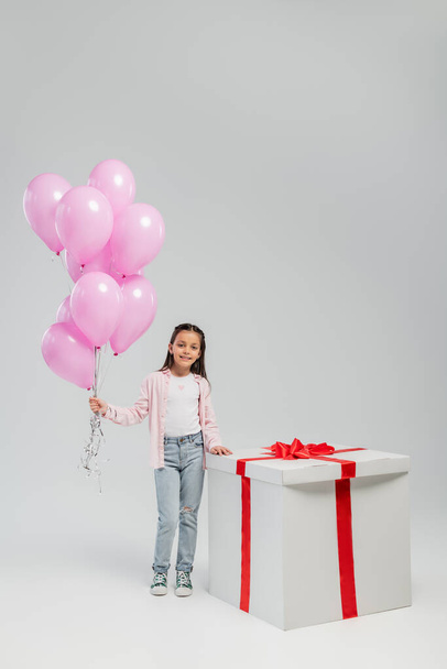 Longitud completa de niño preadolescente despreocupado en ropa casual mirando a la cámara mientras sostiene globos rosados cerca de regalo grande durante el día de protección infantil sobre fondo gris - Foto, imagen