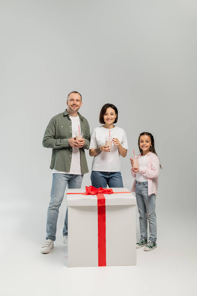 Positive family with preteen daughter holding milkshakes and looking at camera near big gift box during child protection day celebration on grey background - Foto, Bild