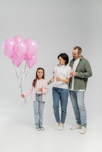 Glimlachende ouders met milkshakes en kijken naar een kleine dochter met ballonnen tijdens internationale kinderfeestjes op grijze achtergrond - Foto, afbeelding