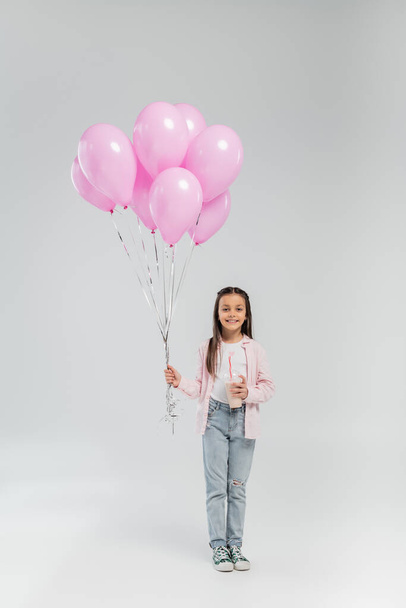 Longitud completa de la niña preadolescente sonriente en ropa casual sosteniendo globos rosados y batido mientras mira a la cámara durante la celebración del día de protección infantil sobre fondo gris - Foto, imagen
