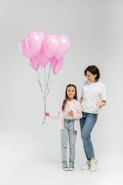 Full length of smiling mother in casual clothes holding milkshake and hugging preteen daughter with pink balloons during child protection day on grey background - Fotoğraf, Görsel