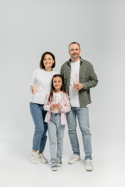 Full length of smiling family with preteen daughter in casual clothes holding milkshakes in plastic cups and looking at camera during children day celebration on grey background - Foto, Imagen