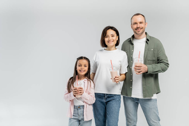 Parents positifs en vêtements décontractés regardant la caméra près de la fille avec milkshake dans une tasse en plastique lors de la célébration de la journée internationale des enfants isolé sur gris avec espace de copie - Photo, image