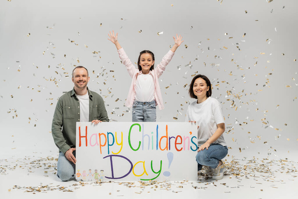 Excited preteen girl in casual clothes waving hands at camera while standing near parent holding placard with happy children's day lettering and under falling festive confetti on grey background - Фото, изображение