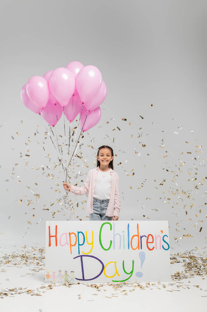 Cheerful preteen kid in casual clothes looking at camera while holding pink balloons near placard with happy children's day lettering and under falling festive confetti on grey background - Photo, Image