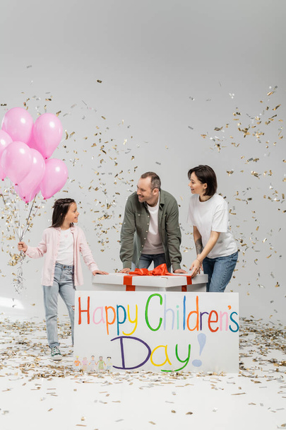 Parents positifs regardant ensemble la fille préadolescente avec des ballons roses près de grand cadeau, pancarte avec le lettrage heureux de la journée des enfants et des confettis tombant pendant la célébration sur fond gris - Photo, image