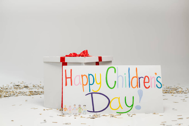 Placard con scritte colorate felici per bambini vicino a una grande confezione regalo con fiocco su sfondo grigio con coriandoli festivi  - Foto, immagini