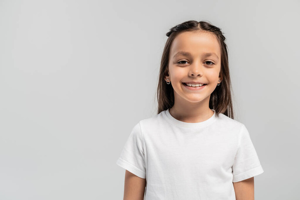 Retrato de pré-adolescente sorridente e menina morena em camiseta branca olhando para a câmera enquanto celebra o dia global de proteção infantil isolado em cinza com espaço de cópia - Foto, Imagem