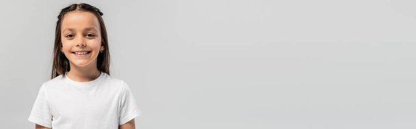 Portrét bezstarostné brunetky předpuberťačky v bílém tričku při pohledu do kamery při mezinárodní oslavě dne ochrany dětí izolované na šedi s kopírovacím prostorem, banner  - Fotografie, Obrázek