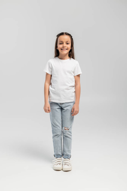 Full length of cheerful and preteen girl in white t-shirt and jeans looking at camera while celebrating global child protection day and standing on grey background - Fotografie, Obrázek