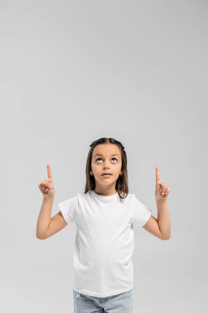 Preadolescente enfocada en camiseta blanca y jeans apuntando con los dedos y mirando hacia arriba durante la celebración del Día Internacional de la Protección del Niño aislada en gris con espacio para copiar  - Foto, Imagen