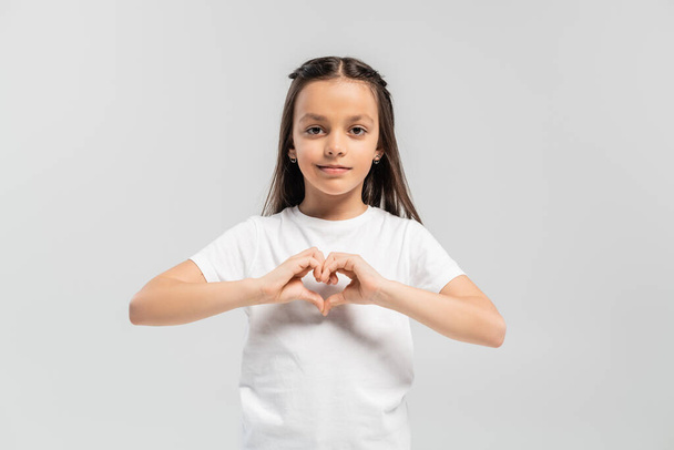 happy preteen girl with long brunette hair standing in white t-shirt and showing heart gesture with hands isolated on grey background, child protection day holiday  - Photo, Image