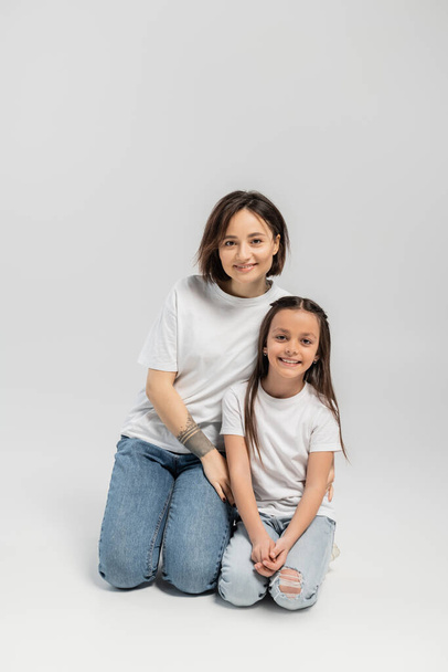 happy mother with tattoo on hand and short hair hugging preteen brunette daughter while sitting together in white t-shirts and blue denim jeans on grey background, child protection day  - Photo, image