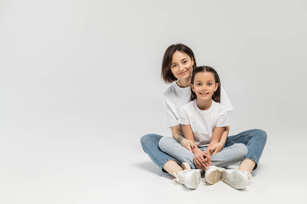 positive mother with tattoo and short hair hugging preteen brunette daughter while sitting together in white t-shirts and blue denim jeans on grey background, child protection day  - Foto, immagini