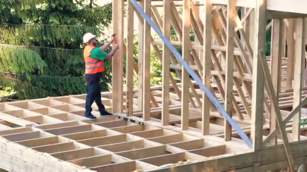Carpenter constructing two-story wooden frame house. Bearded man in glasses hammering nails into structure while wearing protective helmet, construction vest. Concept of modern ecological construction - Footage, Video