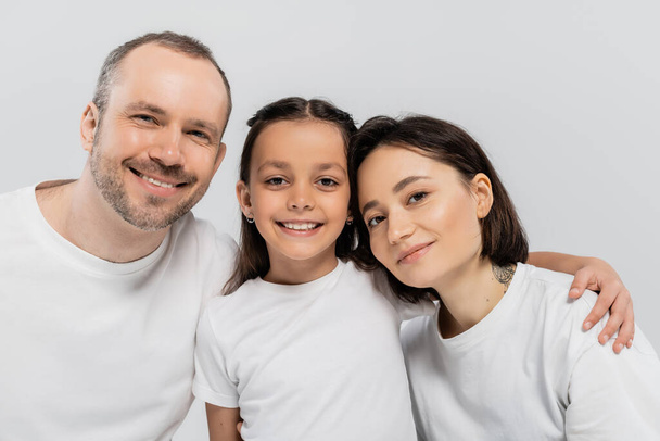 portrait of happy family in white t-shirts looking at camera on grey background, Child protection day, cheerful father and mother with short hair embracing preteen daughter  - Фото, изображение