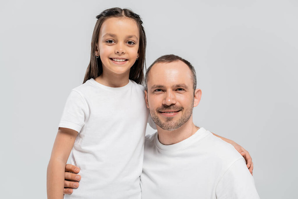 unshaved and cheerful father with bristle hugging happy preteen daughter with long brunette hair while posing in white t-shirts and looking at camera on grey background, Child protection day  - Fotografie, Obrázek