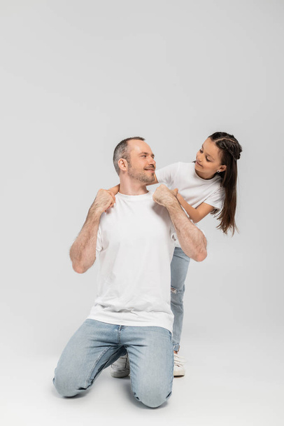 happy preteen girl with long brunette hair leaning on unshaved and cheerful father with bristle while posing in white t-shirts and denim jeans while looking at each other on grey background, Child protection day  - Фото, изображение