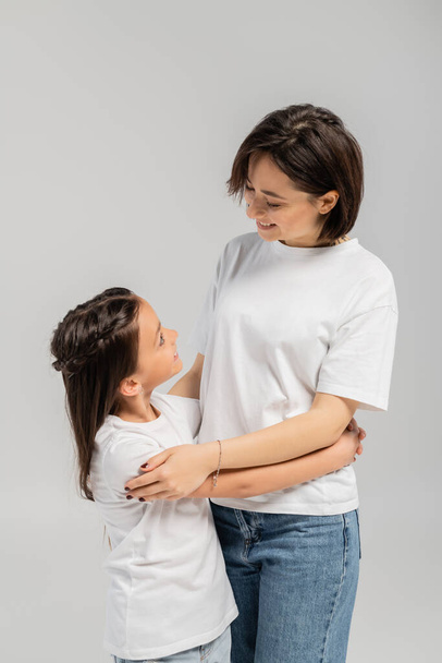 happy mother with short hair and tattoo on hand hugging brunette daughter while standing together in white t-shirts and blue denim jeans on grey background, International child protection day  - Photo, Image