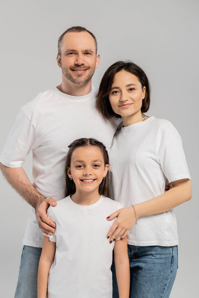 portrait of happy family in white t-shirts looking at camera on grey background, International child protection day, father and mother with tattoo embracing preteen brunette daughter  - Fotografie, Obrázek