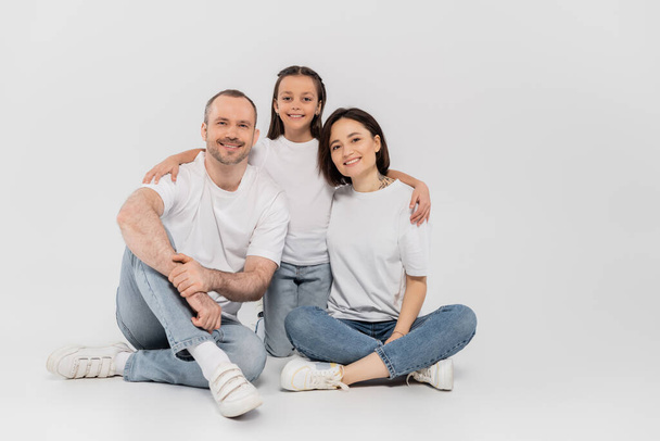 stylish family in white t-shirts and blue denim jeans looking at camera and sitting together on grey background, International child protection day, preteen daughter hugging happy parents  - Foto, Imagem